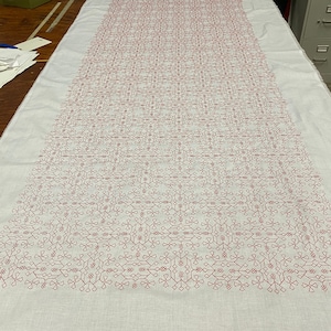 May include: A white fabric tablecloth with a repeating geometric pattern embroidered in a dark red color.
