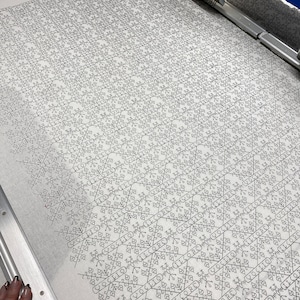 May include: A close-up of a white fabric with a black geometric pattern. The pattern is made up of repeating squares and triangles. The fabric is being embroidered by a machine.