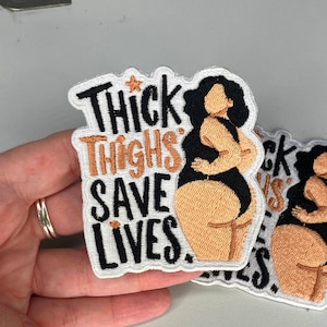 May include: White embroidered patch with a black outline. The patch features a cartoon of a woman with brown skin and black hair, wearing a black swimsuit. The text "Thick Thighs Save Lives." is embroidered in black and brown.