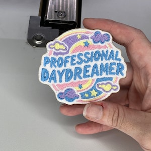 Peut inclure: Un patch brodé blanc avec un arc-en-ciel bleu, rose et jaune, des nuages et des étoiles. Le texte "PROFESSIONAL DAYDREAMER" est brodé en bleu.