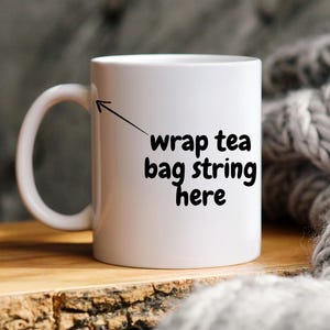 May include: A white ceramic mug with a curved handle. The mug has the text "wrap tea bag string here" in black. The mug is on a wooden surface, with a grey knitted blanket in the background.