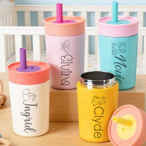 Puede incluir: Una colecci&oacute;n de vasos con boquilla personalizados para beb&eacute;s en varios colores, incluyendo blanco, rosa y amarillo. Cada vaso presenta un nombre y una imagen decorativa. Los vasos tienen tapas y pajitas coloridas, y se exhiben sobre una superficie de madera.