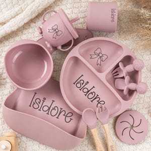 May include: A collection of pink baby feeding essentials, including a divided plate, bowl, bib, cup, and utensils. The set features a bow design and the name "Isidore" in script. The utensils have wooden handles.