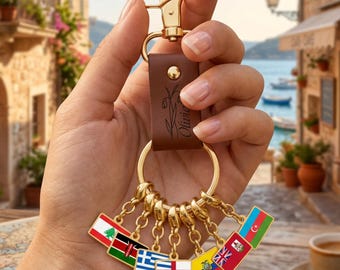 May include: A brown leather keyring featuring a gold-tone metal lobster clasp and a ring holding seven small rectangular charms, each displaying a different national flag in vibrant enamel colours. The leather strap is embossed with a floral design.