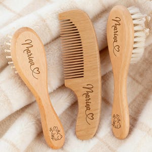 May include: Three wooden baby grooming tools: a brush, a comb, and a brush with soft bristles. Each item is engraved with the name "Marina" and a heart symbol. A small bunny illustration is also engraved on the handles. The items are made of light-colored wood.