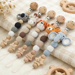 Puede incluir: Pinzas para chupete de bebé con diseños de animales de ganchillo, incluyendo un koala, una jirafa, un zorro y un elefante. Las pinzas tienen cuentas de madera y piezas de madera en forma de estrella con nombres grabados. La paleta de colores incluye tonos neutros y pastel.