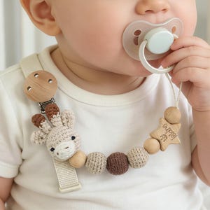 Op de afbeelding: Een baby met een witte romper en een fopspeen. De fopspeen is bevestigd aan een houten en gehaakte fopspeenclip met een giraffendesign, houten kralen en een ster met de naam "Isidre" gegraveerd.