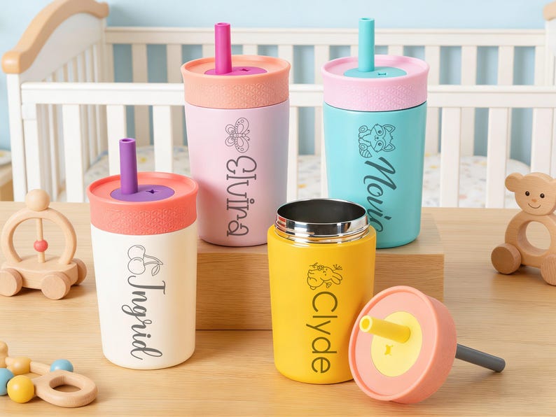 Puede incluir: Una colecci&oacute;n de vasos con boquilla personalizados para beb&eacute;s en varios colores, incluyendo blanco, rosa y amarillo. Cada vaso presenta un nombre y una imagen decorativa. Los vasos tienen tapas y pajitas coloridas, y se exhiben sobre una superficie de madera.