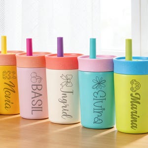 Puede incluir: Un conjunto de vasos aislados de colores con pajitas. Cada vaso presenta una tapa y una pajita de diferente color, con nombres y gr&aacute;ficos decorativos como un conejito, cerezas, un lazo, una flor y un coche. Los vasos est&aacute;n colocados sobre una superficie de madera.