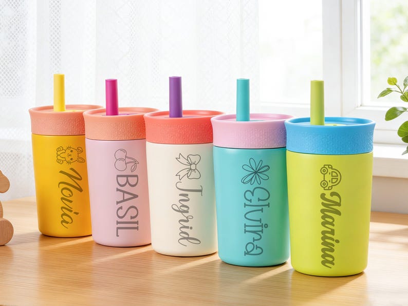 Puede incluir: Un conjunto de vasos aislados de colores con pajitas. Cada vaso presenta una tapa y una pajita de diferente color, con nombres y gr&aacute;ficos decorativos como un conejito, cerezas, un lazo, una flor y un coche. Los vasos est&aacute;n colocados sobre una superficie de madera.