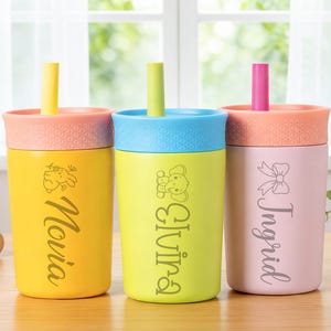 Puede incluir: Tres vasos personalizados con pajitas. Los vasos son amarillos, verdes y rosas, cada uno con una tapa y pajita de diferente color. Los nombres "Novia", "Olivia" e "Ingrid" est&aacute;n impresos en los vasos, con gr&aacute;ficos decorativos.