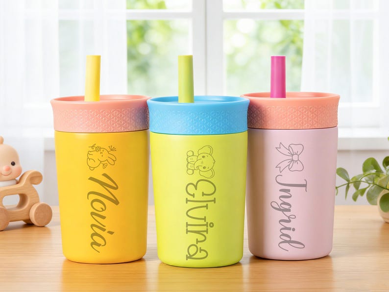 Puede incluir: Tres vasos personalizados con pajitas. Los vasos son amarillos, verdes y rosas, cada uno con una tapa y pajita de diferente color. Los nombres "Novia", "Olivia" e "Ingrid" est&aacute;n impresos en los vasos, con gr&aacute;ficos decorativos.