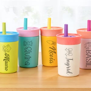 Puede incluir: Cinco vasos aislados de colores con tapas y pajitas. Cada vaso es de un color diferente, incluyendo amarillo, turquesa y rosa. Los vasos tienen nombres y peque&ntilde;as ilustraciones impresas, como un mono, una jirafa, una zanahoria, un lazo y un unicornio.