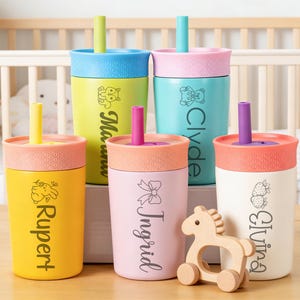 Puede incluir: Una colecci&oacute;n de coloridas tazas para beb&eacute;s con tapas y pajitas. Cada taza es de un color diferente, incluyendo amarillo, rosa, azul y blanco, y presenta un nombre y un peque&ntilde;o gr&aacute;fico. Un caballo de juguete de madera est&aacute; al lado de las tazas.