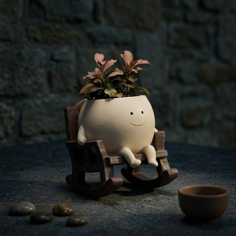 Alt Text: A whimsical cream-colored round face planter with a simple painted smile, sitting relaxed in a miniature brown wooden rocking chair. The cute character pot has small legs dangling down and is holding a vibrant pink and green fittonia nerve plant on a rustic wooden table.