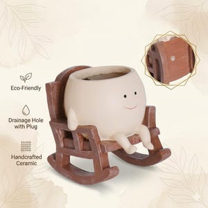 Alt Text: A whimsical cream-colored round face planter with a simple painted smile, sitting relaxed in a miniature brown wooden rocking chair. The cute character pot has small legs dangling down and is holding a vibrant pink and green fittonia nerve plant on a rustic wooden table.