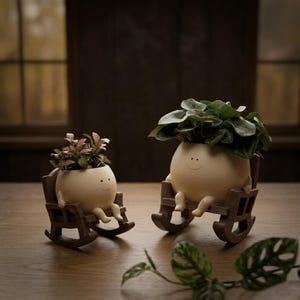 A whimsical cream-colored round face planter with a simple painted smile, sitting relaxed in a miniature brown wooden rocking chair. The cute character pot has small legs dangling down and is holding a vibrant pink and green fittonia nerve plant on a rustic wooden table.