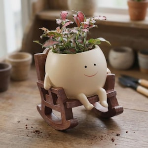 May include: A cream-colored ceramic planter shaped like a round character with a simple smiling face, seated in a small brown wooden rocking chair. The planter holds a small plant with variegated green and pink leaves, resting on a wooden surface.