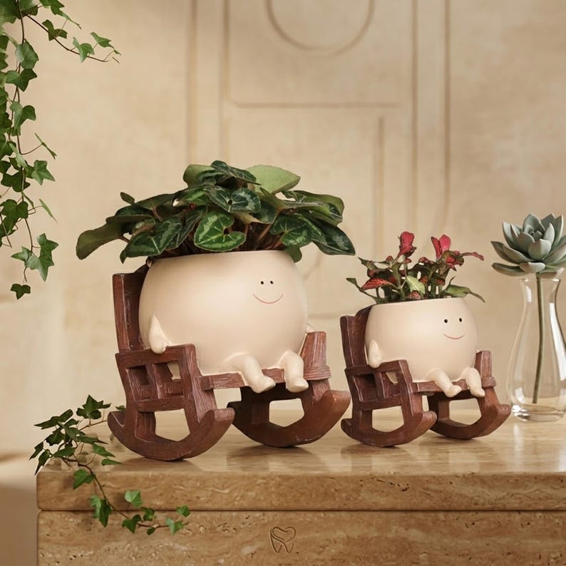 Alt Text: A whimsical cream-colored round face planter with a simple painted smile, sitting relaxed in a miniature brown wooden rocking chair. The cute character pot has small legs dangling down and is holding a vibrant pink and green fittonia nerve plant on a rustic wooden table.