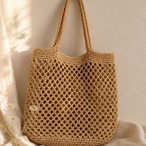 May include: A tan crochet tote bag featuring an open diamond-shaped mesh pattern throughout the body and two integrated shoulder straps. The bag is constructed from a textured fiber material with a consistent light brown color and a structured base.