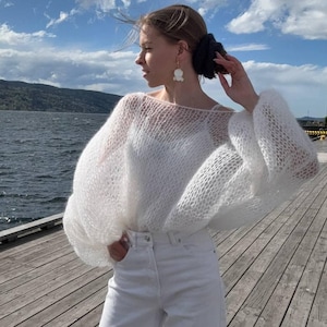 Op de afbeelding: Een vrouw draagt een witte, open geweven trui met volumineuze mouwen en een witte jeans. De trui heeft een boothals en een losse pasvorm. De achtergrond omvat een water, een houten pier en een blauwe lucht met wolken.