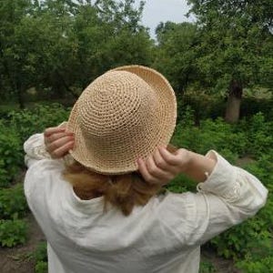 Puede incluir: Un sombrero de color beige claro con copa redonda hecho de material de paja trenzada. El sombrero presenta un ala corta ligeramente levantada y un patrón circular texturizado en toda la copa, mostrando un color natural y un diseño simple y estructurado.