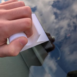 Könnte beinhalten: Ein weißer Kunststoff-Abzieher mit rundem Griff wird auf einer Glasoberfläche verwendet. Das Glas reflektiert einen bewölkten Himmel. Der Abzieher ist für die Reinigung von Fenstern und anderen glatten Oberflächen konzipiert.