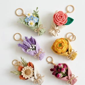 May include: A collection of handmade crochet bouquet keychains. Each keychain features a miniature bouquet of colorful flowers, including roses, daisies, and lavender, wrapped in a crocheted paper. The keychains have gold-colored metal rings.