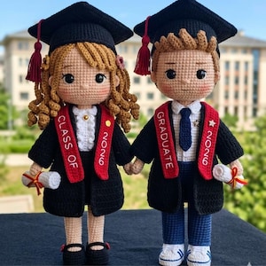 Puede incluir: Dos muñecos de graduación de ganchillo, una niña y un niño, tomados de la mano. Llevan vestidos de graduación negros, gorras con borlas y estolas rojas que dicen "Class of 2026" y "Graduate". Cada muñeco sostiene un diploma.