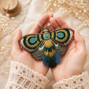 Peut inclure: Broche en forme de papillon de nuit faite à la main, avec des ailes en crochet vert d'eau, vert et or, bordées de marron et une queue en plumes. La broche présente une tête en métal détaillée.
