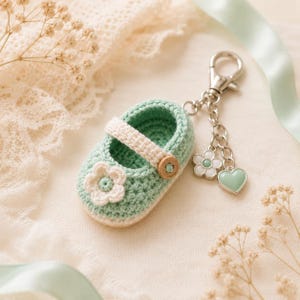 May include: A crocheted baby shoe keychain in mint green and cream. The shoe features a flower accent and a wooden button. A silver chain holds the shoe, along with a flower and heart charm. The background is a soft, neutral color.
