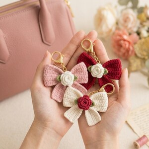 May include: Three handmade crochet bow keychains in pink, red, and cream, each adorned with a small floral accent. The keychains have gold-colored clasps. The bows are held in open hands, with a pink purse in the background.