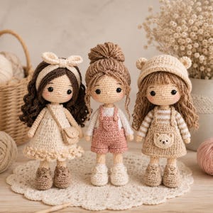 May include: Three handmade crochet dolls standing side by side. The left doll has dark brown curly hair with a cream headband and dress. The center doll has light brown hair in a bun and wears pink overalls. The right doll wears a bear-ear hat.