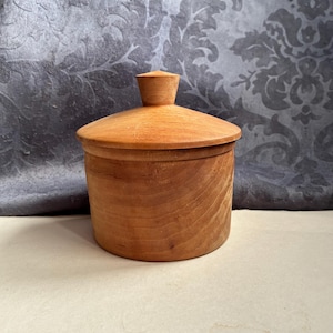 May include: A cylindrical wooden container with a smooth, light brown finish and visible natural wood grain patterns. The container features a matching circular lid with a small, tapered knob handle centered on top for easy removal and access.