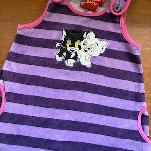 Puede incluir: Vestido de bebé a rayas moradas y lavanda con una aplicación de gato. El vestido tiene ribetes rosas y detalles de botones en los hombros. La etiqueta dice "Bamse by Linden".