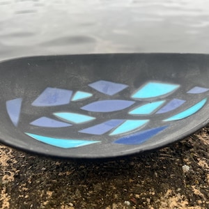 May include: A black ceramic dish with a unique design. The dish features a series of blue and turquoise diamond-shaped patterns. The dish is oval-shaped and sits on a textured, brown surface.