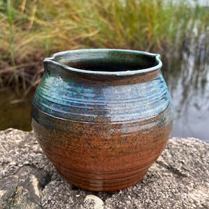 May include: A round ceramic pot with a wide opening and a small spout. The lower portion features a warm, earthy brown glaze, while the upper section transitions into a deep, glossy teal blue. The surface has horizontal ridges and a smooth finish.