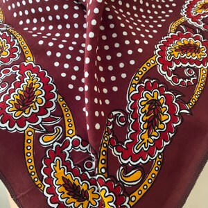 Peut inclure: Un foulard carré présentant un fond bordeaux profond avec un motif à pois blancs sur la partie supérieure. La bordure inférieure affiche un imprimé cachemire vibrant dans des tons de rouge, jaune et blanc avec des contours noirs.