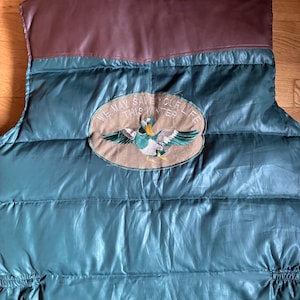 May include: A dark green quilted puffer vest with a brown yoke. Centered on the back is an oval patch featuring an embroidered mallard duck and the text "WE MAY SAVE YOUR LIFE THIS WINTER" stitched in a circular pattern around the duck.