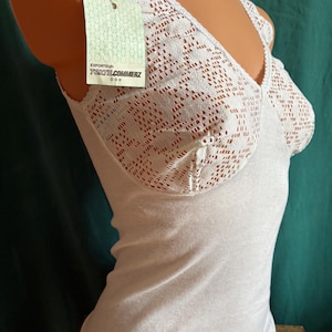 May include: A white sleeveless top featuring a V-neckline with intricate lace detailing on the upper bodice. A light green rectangular tag is attached to the shoulder strap, displaying the text EXPORTEUR: TEXTILCOMMERZ DDR in black lettering.