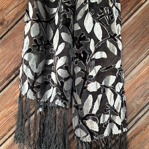 May include: A black and gray scarf with a leaf pattern and fringe. The scarf features a repeating design of leaves in shades of gray and white against a black background. The edges are finished with long, black fringe.