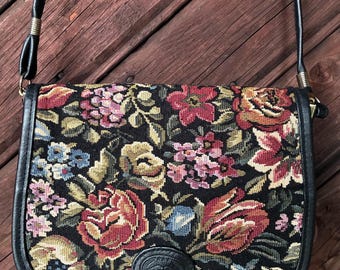 May include: A saddle-shaped crossbody bag featuring a black background with a multi-colored floral tapestry pattern. The bag has black leather trim, a long black shoulder strap, and a small black leather oval patch embossed with a logo at the bottom.