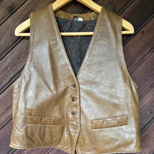 May include: A tan leather vest featuring a V-neckline, four front buttons, and two horizontal welt pockets. The interior is lined with a dark fabric displaying a multi-colored paisley pattern. The garment is displayed on a wooden hanger.