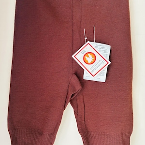 Puede incluir: Leggings de punto en color marrón oscuro sólido con cintura acanalada y puños acanalados en los tobillos. Una etiqueta de producto rectangular blanca con bordes rojos y un logotipo circular que muestra dos figuras está sujeta al frente.