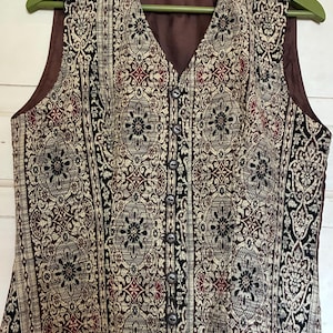 Op de afbeelding: Een mouwloos vest met een complex patroon in tapijstijl in de kleuren zwart, crème en donkerrood. Het kledingstuk heeft een V-hals, een knoopsluiting aan de voorzijde met zeven donkere knopen en een effen bruine voering met Kellermann-label.