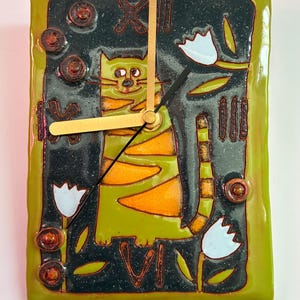 Può includere: Un orologio da parete rettangolare con bordo verde e sfondo nero. Il quadrante presenta un gatto dei cartoni animati con strisce arancioni e verdi, lancette dorate e accenti floreali bianchi. L'orologio misura circa 20 cm per 15 cm.