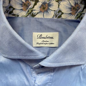 Könnte beinhalten: Nahaufnahme eines hellblauen Hemdes mit floral gemustertem Kragen. Das Hemd hat einen Button-Down-Kragen und ein Etikett mit der Aufschrift "Stenströms. Slimline Twofold super cotton."