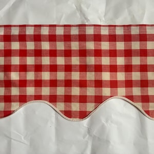 Op de afbeelding: Een raamvalance met een klassiek rood-wit gingham ruitpatroon. De stof heeft een geschulpte onderrand afgewerkt met een dunne, lichtgekleurde bies, wat zorgt voor een herhalend gebogen ontwerp langs de onderste rand van het textiel.