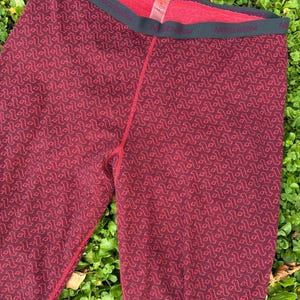 May include: Burgundy leggings featuring a repeating geometric pattern in a darker shade of red. The garment has a dark gray elastic waistband with the brand name Norheim printed in a repeating pattern. The interior lining is a bright red color.