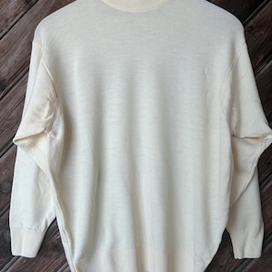 Puede incluir: Jersey de cuello alto color crema con mangas largas. El jersey está hecho de un material de punto suave y presenta detalles acanalados en el cuello, los puños y el dobladillo. El jersey está colgado en una percha de madera sobre un fondo de madera.